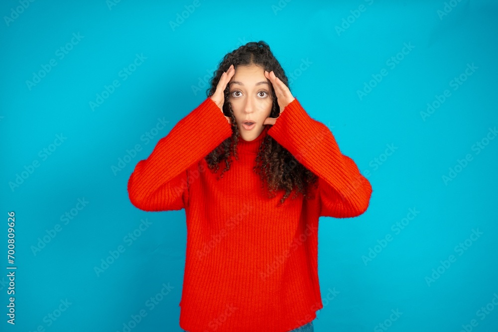 Beautiful teen girl wearing knitted red sweater over blue background with scared expression, keeps hands on head, jaw dropped, has terrific expression. Omg concept Beautiful teen girl wearing knitted red sweater over blue background with scared expression, keeps hands on head, jaw dropped, has terrific expression. Omg concept