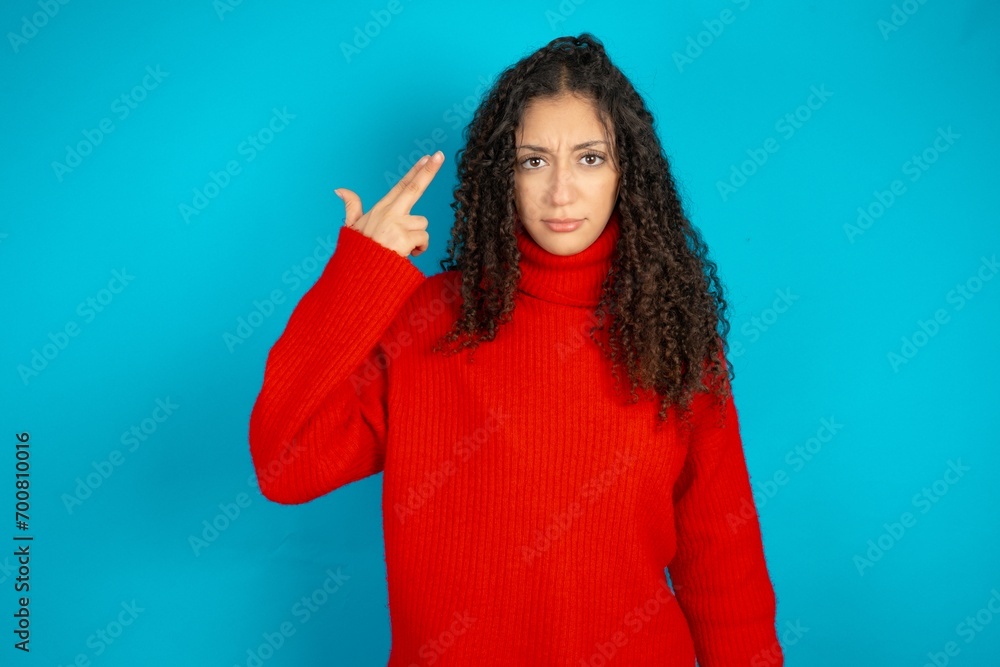 Unhappy Beautiful teen girl wearing knitted red sweater over blue background makes suicide gesture and imitates gun with hand, curves lips, keeps two fingers on temple
