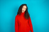 Beautiful teen girl wearing knitted red sweater over blue background winking looking at the camera with sexy expression, cheerful and happy face.