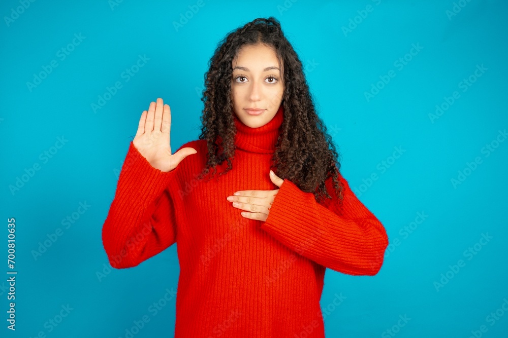 Beautiful teen girl wearing knitted red sweater over blue background Swearing with hand on chest and open palm, making a loyalty promise oath Beautiful teen girl wearing knitted red sweater over blue background Swearing with hand on chest and open palm, making a loyalty promise oath