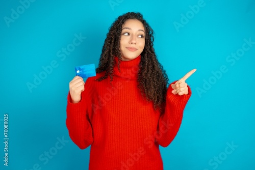 Curious smiling beautiful teen girl wearing red knitted sweater showing plastic bank showing finger copyspace photo