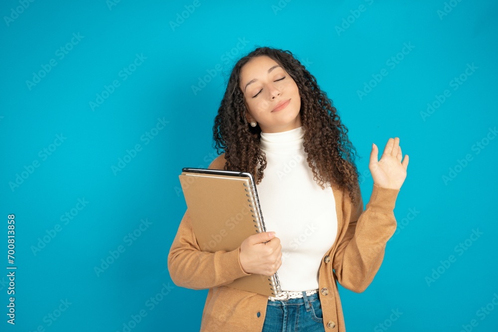 Fototapeta premium Overjoyed successful beautiful arab student carries notebooks raises palm and closes eyes in joy being entertained by friends