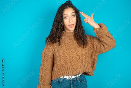 Obraz na plátně Beautiful teen girl wearing knitted sweater over blue background foolishness around shoots in temple with fingers makes suicide gesture