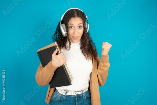 Emotional beautiful caucasian teen girl  exclaims loudly feels like winner raises clenched fists keeps mouth opened wears stereo headphones on ears makes yes gesture, listens favorite music