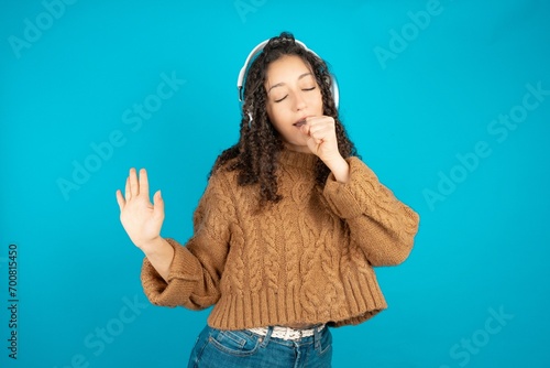 Happy beautiful caucasian teen girl wearing brown sweater sings favourite song keeps hand near mouth as if microphone wears wireless headphones, listens music