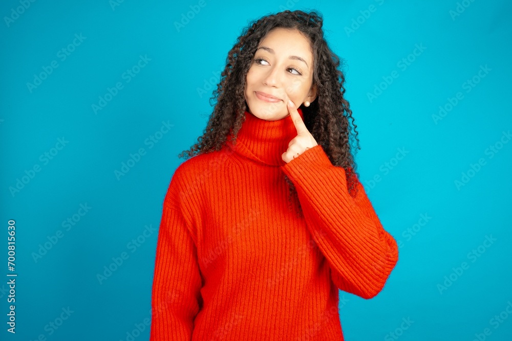 Fototapeta premium Lovely dreamy beautiful teen girl wearing red knitted sweater keeps finger near lips looks aside copy space.