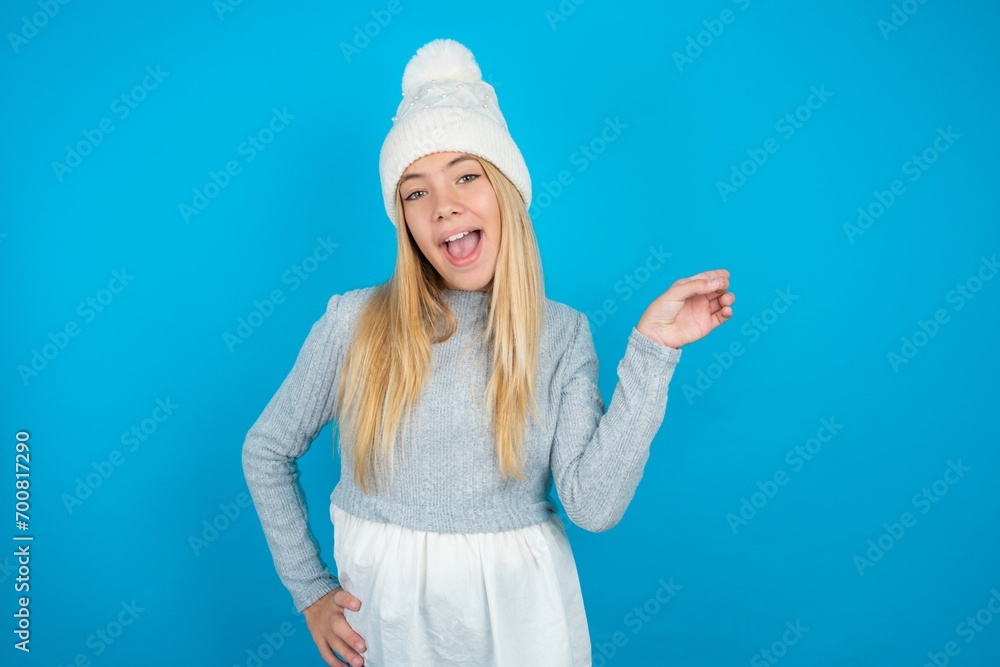 Teen caucasian girl wearing blue knitted sweater and woolly hat holding finger up having idea and posing Teen caucasian girl wearing blue knitted sweater and woolly hat holding finger up having idea and posing