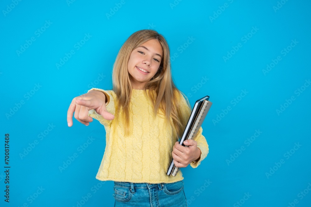 Beautiful kid girl wearing yellow sweater holding notebook pointing at camera with a satisfied, confident, friendly smile, choosing you Beautiful kid girl wearing yellow sweater holding notebook pointing at camera with a satisfied, confident, friendly smile, choosing you