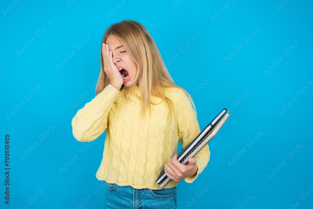 Beautiful kid girl wearing yellow sweater holding notebook Yawning tired covering half face, eye and mouth with hand. Face hurts in pain. Beautiful kid girl wearing yellow sweater holding notebook Yawning tired covering half face, eye and mouth with hand. Face hurts in pain.