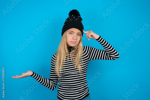 Teen caucasian girl wearing striped sweater and woolly hat confused and annoyed with open palm showing copy space and pointing finger to forehead. Think about it.