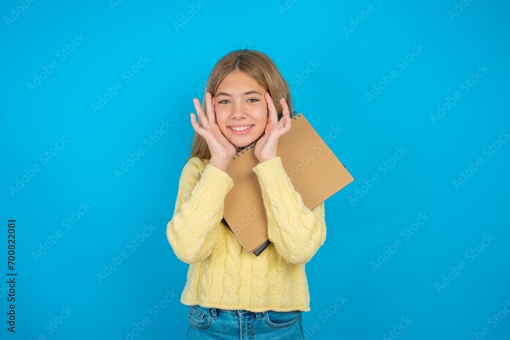 Happy beautiful caucasian teen girl wearing yellow sweater touches both cheeks gently, has tender smile, shows white teeth, gazes positively straightly at camera, Happy beautiful caucasian teen girl wearing yellow sweater touches both cheeks gently, has tender smile, shows white teeth, gazes positively straightly at camera,