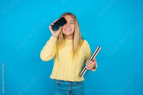 beautiful caucasian teen girl wearing yellow sweater holding modern smartphone covering one eye while smiling