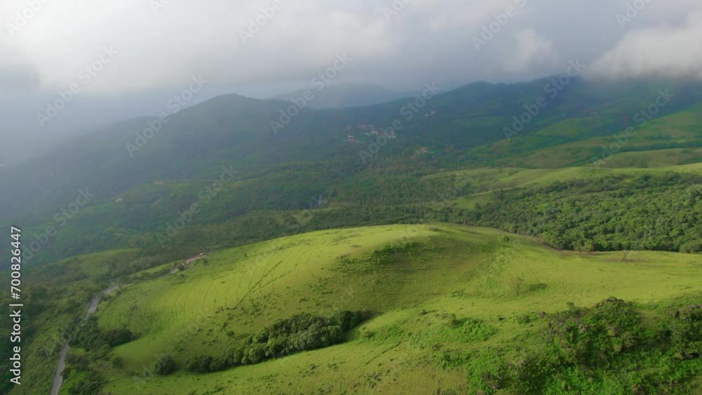 A drone aerial shot of a scenic beautiful hill mound in the foreground with lush green meadows and trees in misty monsoon weather with clouds on top of the mountain range of Indian western ghats. A drone aerial shot of a scenic beautiful hill mound in the foreground with lush green meadows and trees in misty monsoon weather with clouds on top of the mountain range of Indian western ghats.