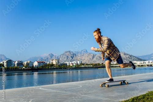 handsome redhaired man with long beard skateboarding near river at sunny summer day