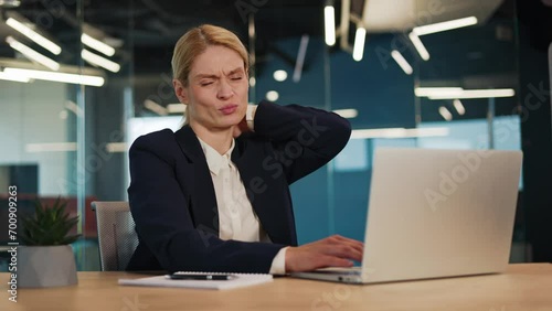 Exhausted businesswoman seated at her desk, enduring intense neck pain and alleviating muscle tension through gentle rubbing. Entrepreneur struggles with spinal tension in office, sedentary lifestyle. photo