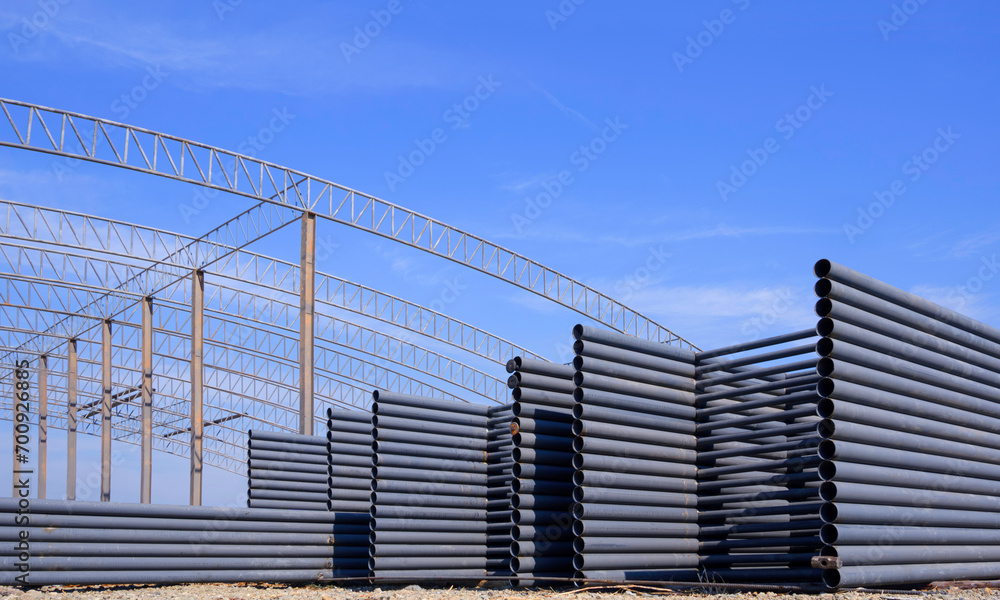 Pile of many metal roof beams for installing on top of large industrial factory building outline structure in construction site area against blue sky background Pile of many metal roof beams for installing on top of large industrial factory building outline structure in construction site area against blue sky background