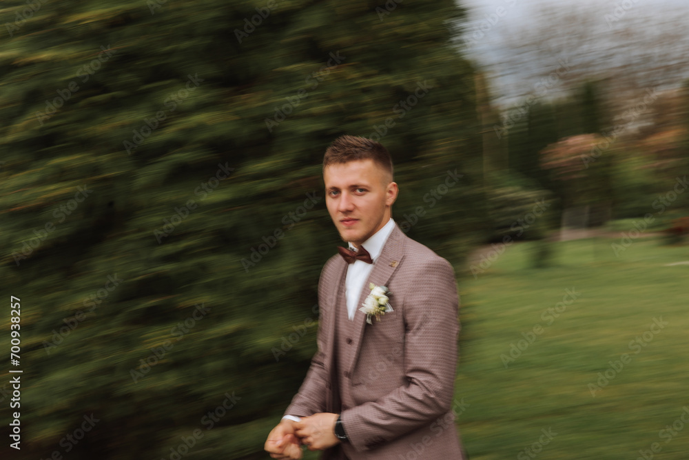The groom in a brown suit adjusts his jacket, poses against the background of green trees. Wedding portrait. The groom in a brown suit adjusts his jacket, poses against the background of green trees. Wedding portrait.