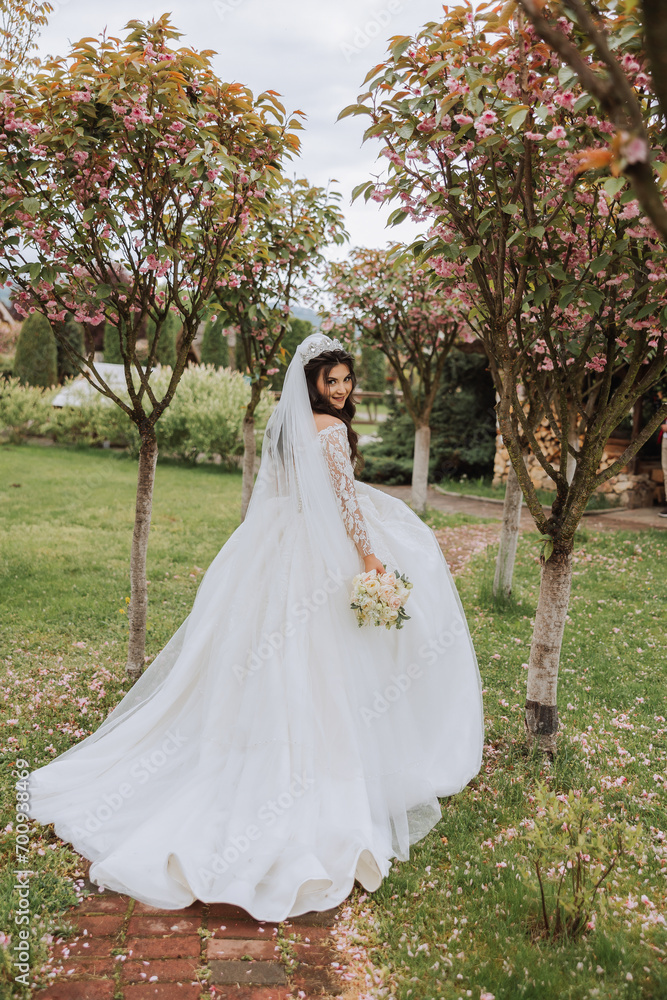 Curly brunette bride in a lush veil and long-sleeved dress poses near cherry blossoms. Spring wedding