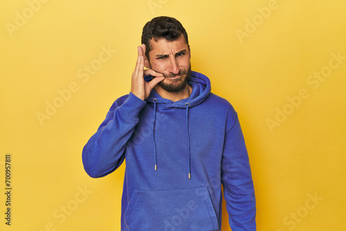 Young Hispanic man on yellow background with fingers on lips keeping a secret.