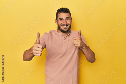 Young Hispanic man on yellow background raising both thumbs up, smiling and confident.