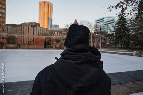 Young man on his back with a big blue buso and black hat looking at a tourist track for ice skating with some Christmas decorations in the background In the center of Calgary Canada  photo