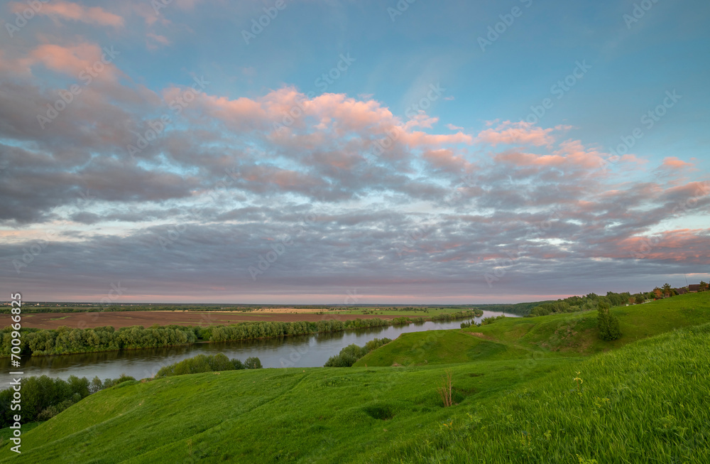 A river against a backdrop of green fields, a breathtaking view from the hill. Colorful sky with clouds during sunset. Landscape in spring. Nature during a bright sunset.