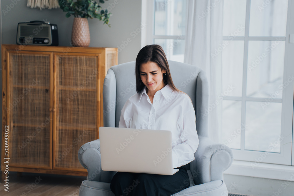 Smiling woman in white shirt using a laptop comfortably seated in a gray armchair, with a bright window and stylish decor Smiling woman in white shirt using a laptop comfortably seated in a gray armchair, with a bright window and stylish decor