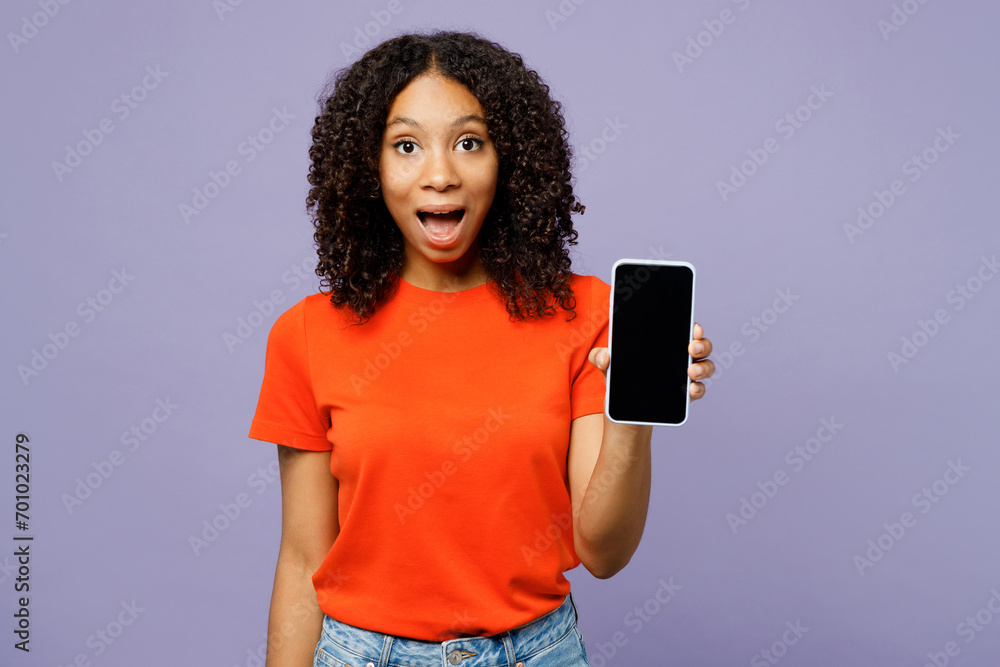 Little kid teen girl of African American ethnicity in orange t-shirt hold in hand use blank screen area mobile cell phone isolated on plain pastel light purple background Childhood lifestyle concept Little kid teen girl of African American ethnicity in orange t-shirt hold in hand use blank screen area mobile cell phone isolated on plain pastel light purple background Childhood lifestyle concept