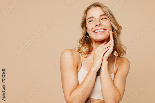 Close up young happy nice lady woman with slim body perfect skin wears nude top bra lingerie stand put hand on face looking aside isolated on plain pastel beige background. Lifestyle diet fit concept.