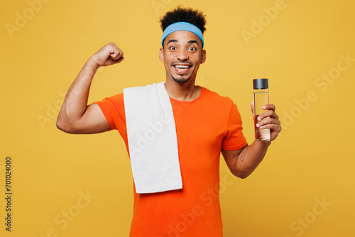 Young fitness trainer instructor sporty man sportsman wear orange t-shirt hold water bottle towel show muscle spend time in home gym isolated on plain yellow background. Workout sport fit abs concept.