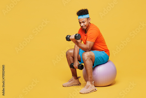 Full body sad young fitness trainer sporty man sportsman wearing orange t-shirt sit on fit ball hold use dumbbells training in home gym isolated on plain yellow background. Workout sport abs concept.