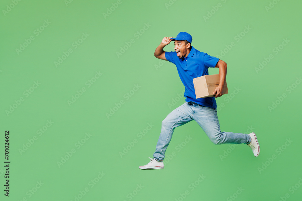 Full body fun delivery guy employee man wear blue cap t-shirt uniform workwear work as dealer courier jump high with blank craft cardboard box run isolated on plain green background. Service concept.