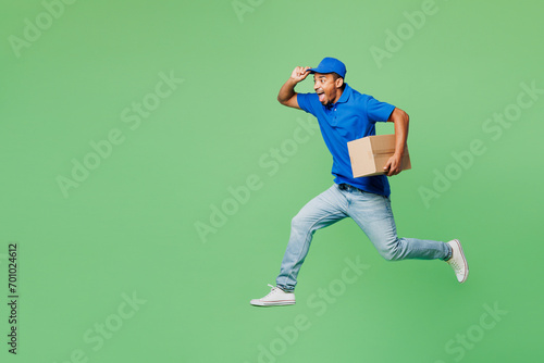 Full body fun delivery guy employee man wear blue cap t-shirt uniform workwear work as dealer courier jump high with blank craft cardboard box run isolated on plain green background. Service concept. photo