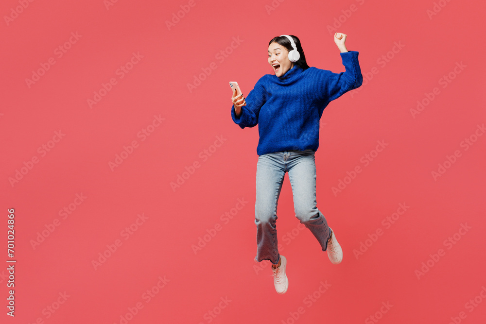 Full body young winner woman of Asian ethnicity she wear blue sweater casual clothes jump high hold use mobile cell phone listen to music in headphones isolated on plain pastel light pink background.