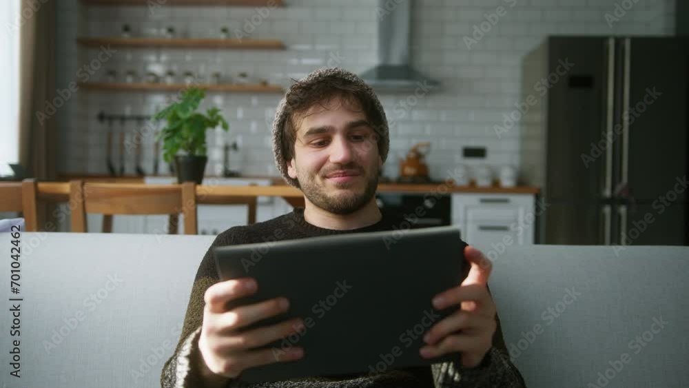Young man with beanie sitting on sofa at home with kitchen background, playing excited concentrated engrossed in interesting mobil game with tablet computer