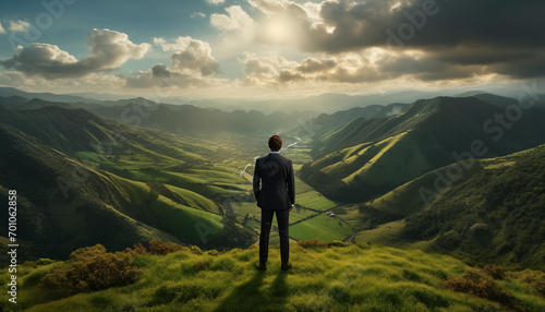 Business man in dark suite standing on green grass in the mountains looking into a scenic valley with light rays and dramatic clouds. Concept of new frontiers and success.  © iconimage