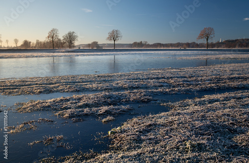 Landscape shot of a field in winter. The field is partially covered with frozen water. Trees reflected on the horizon. The grass is frozen.