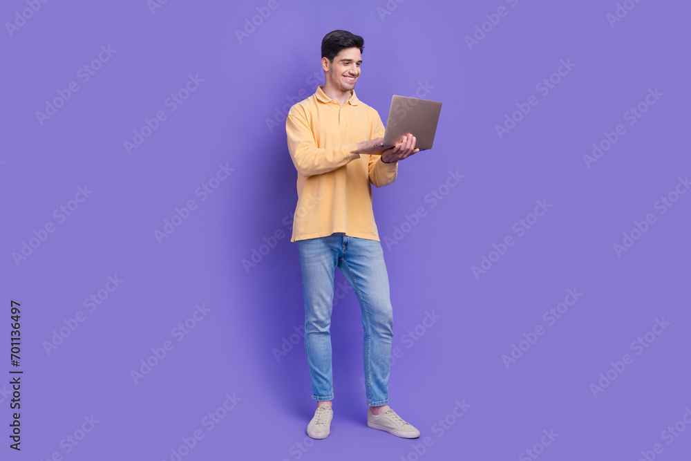 Full size photo of attractive young man hold netbook typing report work wear trendy yellow clothes isolated on violet color background Full size photo of attractive young man hold netbook typing report work wear trendy yellow clothes isolated on violet color background