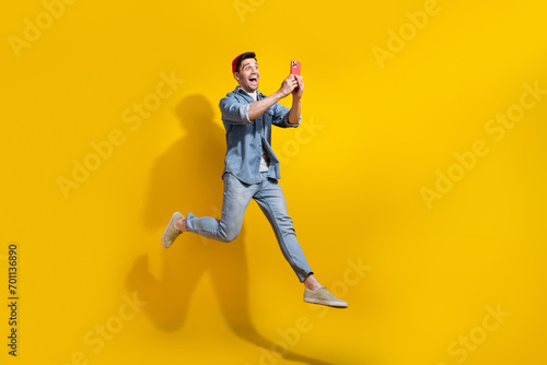 Full length photo of impressed funky man dressed jeans shirt jumping high chatting modern device empty space isolated yellow color background