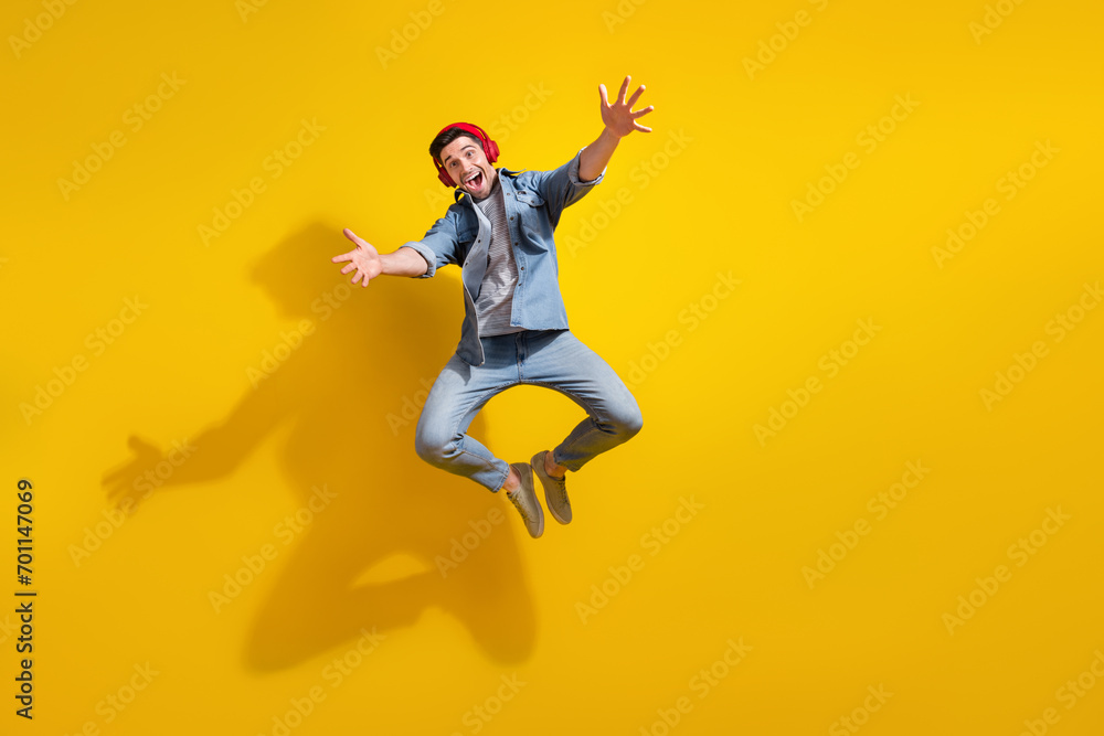 Full length photo of carefree friendly man dressed jeans shirt jumping listening music open arms ready hug you isolated yellow color background