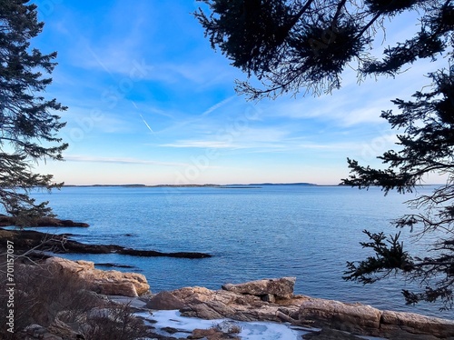 View of water and blue sky framed by leaves of trees in winter, boulders and a bit of snow in foreground. LaVerna Preserve hiking trails, Maine, US. Horizontal.