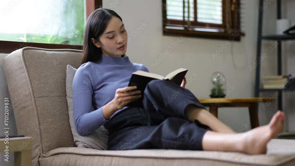 Charming Asian woman is happily reading a book on the sofa in the living room at home. Charming Asian woman is happily reading a book on the sofa in the living room at home.