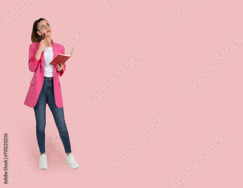 Thoughtful concentrated woman, full body portrait of charming thoughtful concentrated woman. Holding pen and notepad, thinking looking copy space. Isolated pastel pink background. Advertisement idea. © Designerant