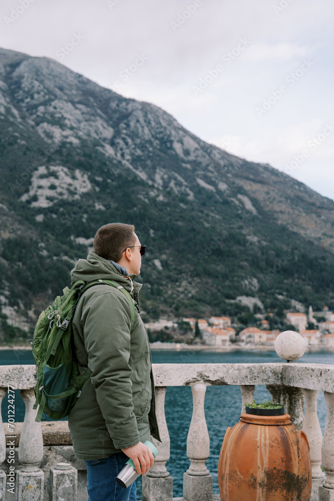 Traveler with a thermos and a backpack stands on the seashore at the railing and looks at the mountains. Side view