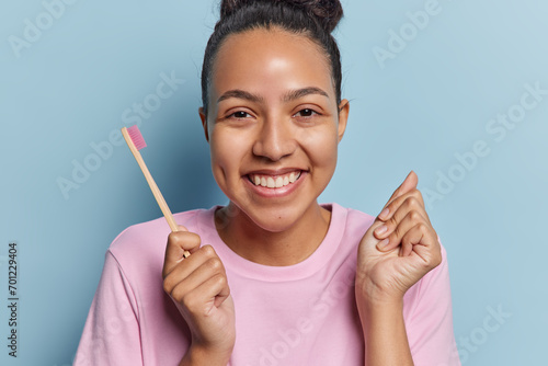 Smiling cheerful Latin woman with dark hair gathered in bun holds wooden toothbrush going to clean teeth smiles broadly clenches fist dressed in casual t shirt isolated over blue background. photo
