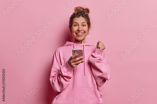 Cheerful overjoyed woman with hair bun clenches fist and rejoices success holds mobile phone in hand let out joyful sigh expresses sense of achievement wears casual sweatshirt isolated on pink wall