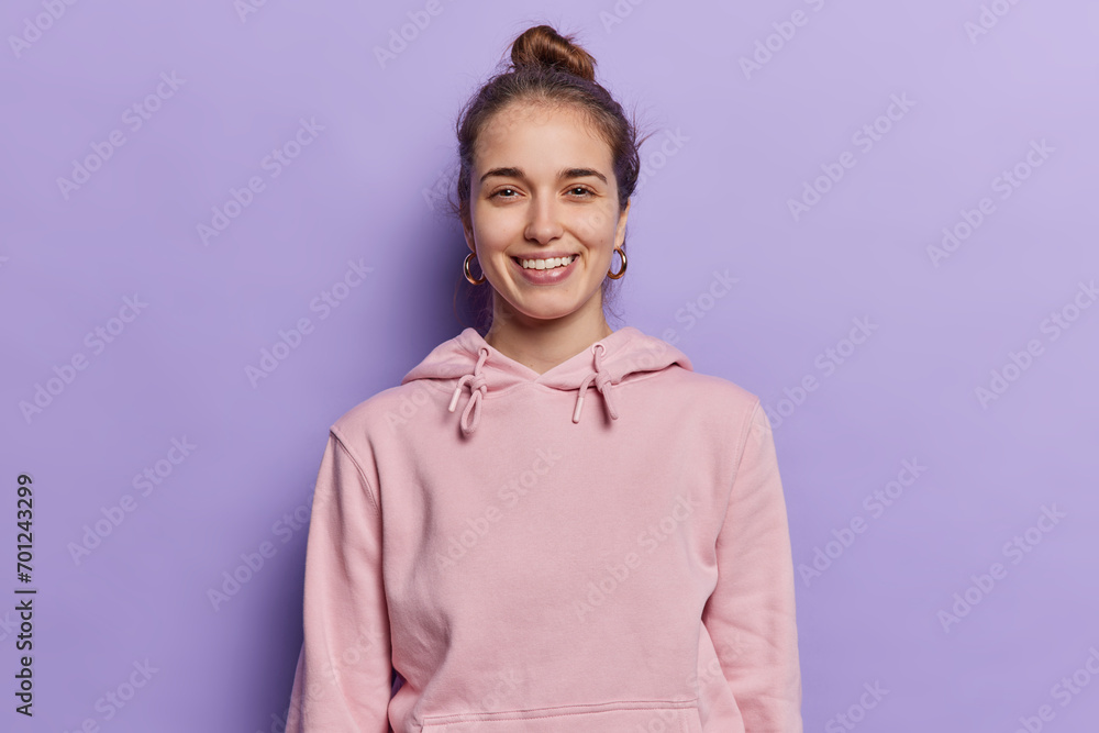 People positive emotions concept. Indoor waist up of young pretty glad smiling European lady standing in centre isolated on purple background wearing pink hoodie keeping hands down looking straight People positive emotions concept. Indoor waist up of young pretty glad smiling European lady standing in centre isolated on purple background wearing pink hoodie keeping hands down looking straight