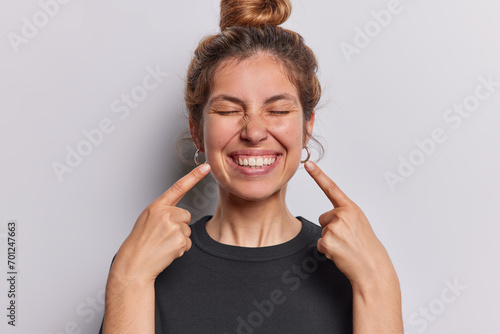 Photo of pleasant looking cheerful woman points index fingers at toothy smile shows wwell cared whie even teeth dressed in casual black t shirt isolated over whtie studio background feels happy