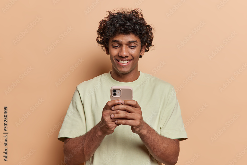 Cheerful handsome young Hindu man holding smartphone watches video and looks at screen chatting online with friend using application dressed in casual t shirt isolated over brown background. Cheerful handsome young Hindu man holding smartphone watches video and looks at screen chatting online with friend using application dressed in casual t shirt isolated over brown background.