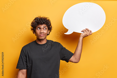 People emotions concept. Indoor photo of young disappointed Hindu male standing in centre isolated on yellow background wearing black t shirt holding and looking at speech bubble with blank space photo